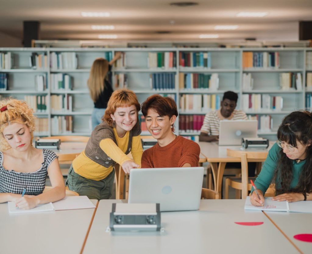 Multiethnic male and female students taking notes and using laptops in library Cheerful young multiracial friends smiling and pointing at laptop screen while preparing for exams near concentrated male and female classmates in modern library