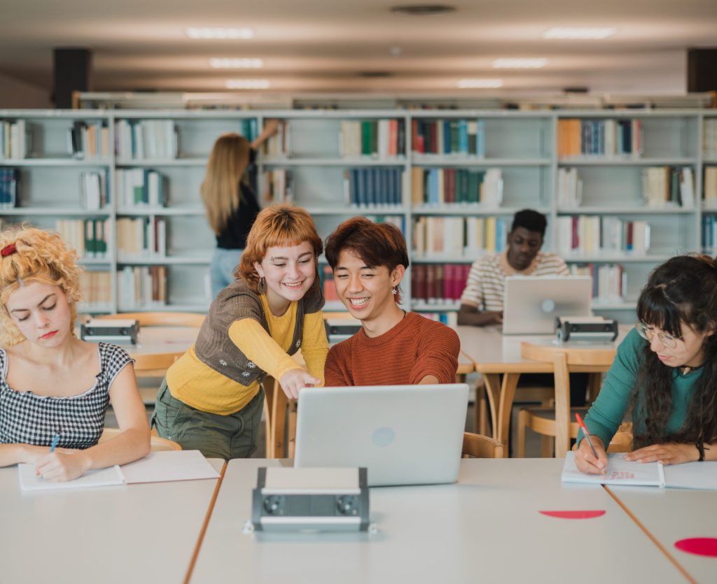 Cheerful young multiracial friends smiling and pointing at laptop screen while preparing for exams near concentrated male and female classmates in modern library