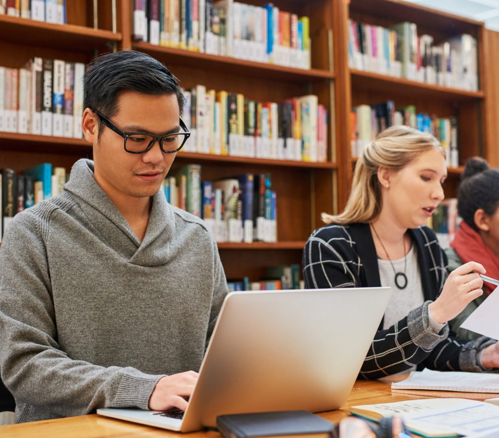 Laptop, typing and man in library for study with university, education and scholarship for growth. Books, online research and college student on campus with learning, knowledge and writing paper.